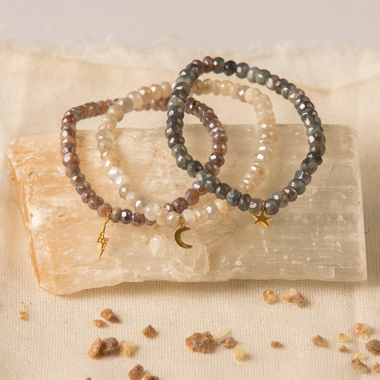Hand-beaded set of three moonstone bracelets with subtle color variations (grey - smoke, brown - taupe, cream - pearl) and gold celestial charms (lightning bolt, moon, star). This stackable moonstone jewelry offers protection, love, and connection to cosmic energy.